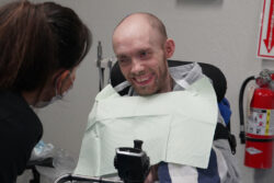 ACRC individual Charles sits in a wheelchair in a dentist office and smiles at the medical staff treating him.
