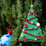An ornament in the shape of a tree with mini ornaments, red ribbon and a star. There's also a blue ball ornament in the background.